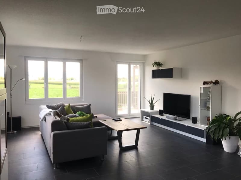 Modern living room with large windows offering a view, featuring a grey sofa with multiple cushions, a wooden coffee table, a flat-screen TV on a sleek entertainment unit, and a floor lamp.