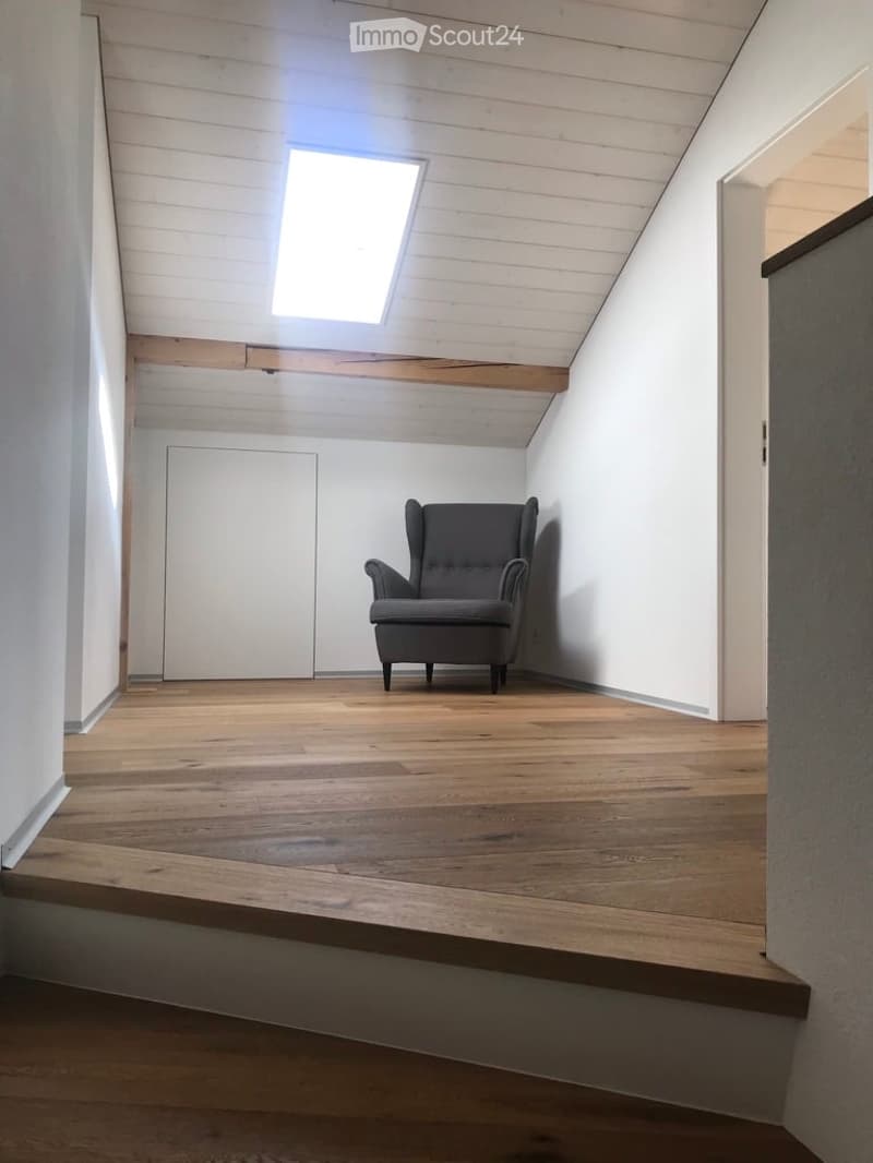 Minimalist room with wooden flooring, white walls, skylight window, and a single grey armchair.