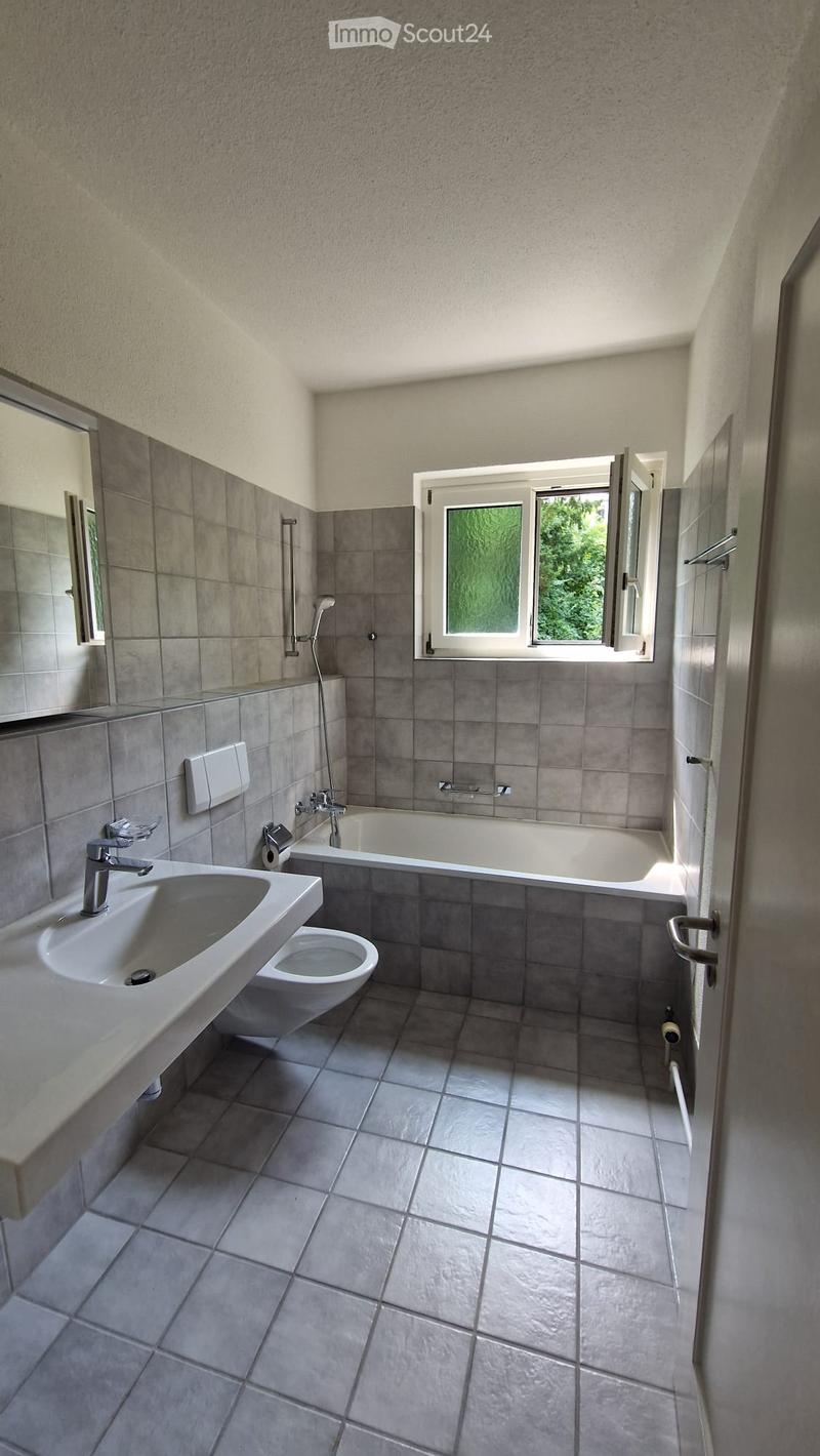 Modern bathroom with grey tiles, a large window, a sink, toilet, bathtub, and a mirror.