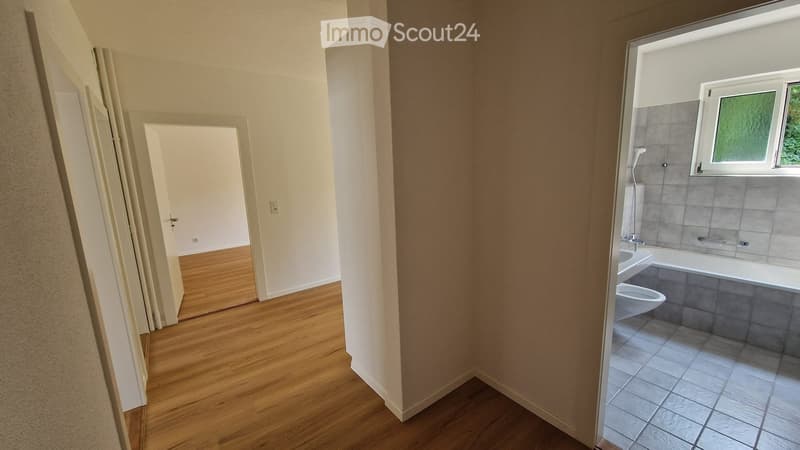 Empty hallway leading to a bathroom with a bathtub, toilet, and tiled walls. The hallway has wooden flooring and white walls.