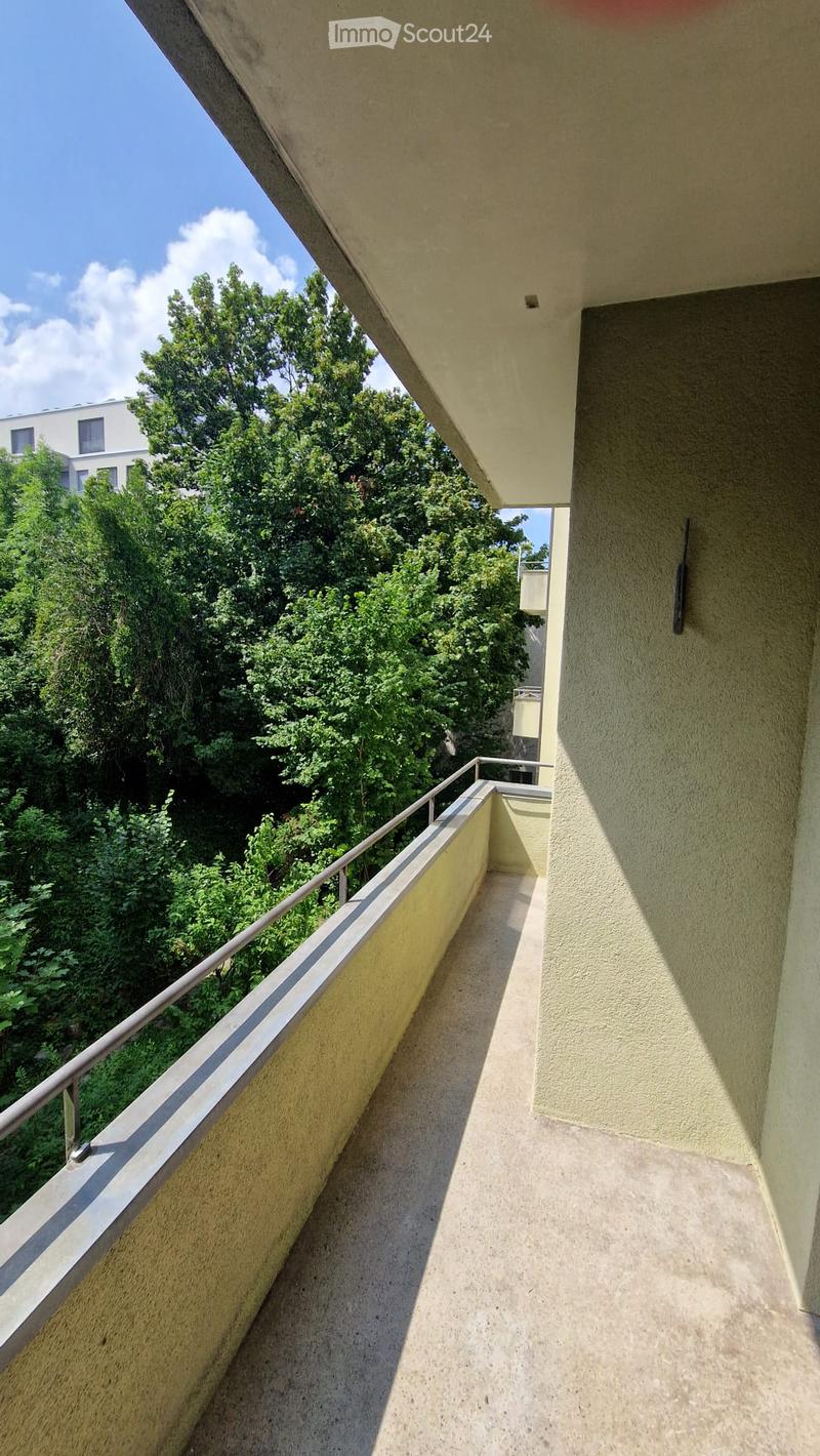 Balcony with metal railing, overlooking a green area with trees and a neighboring building.