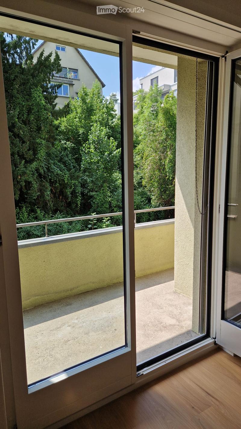Open sliding glass door leading to a balcony with a metal railing, overlooking a green garden area with trees and neighboring buildings.