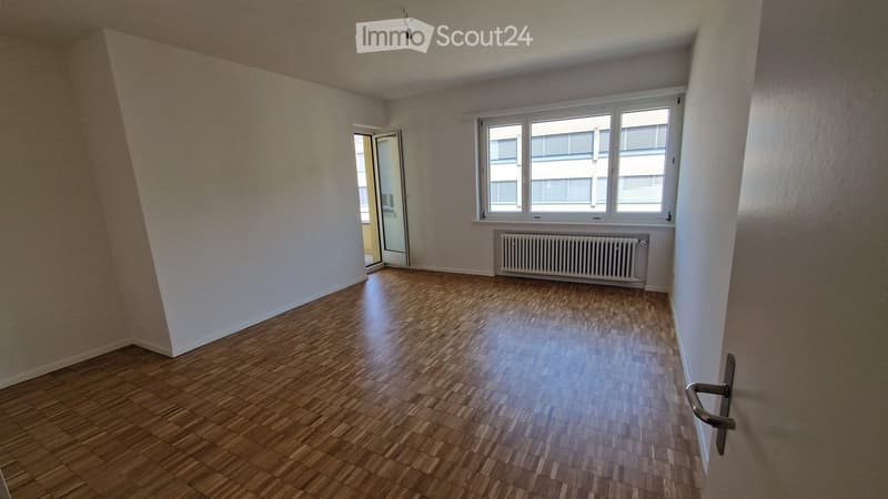 Empty room with wooden flooring, white walls, large windows, and a radiator under the windows.