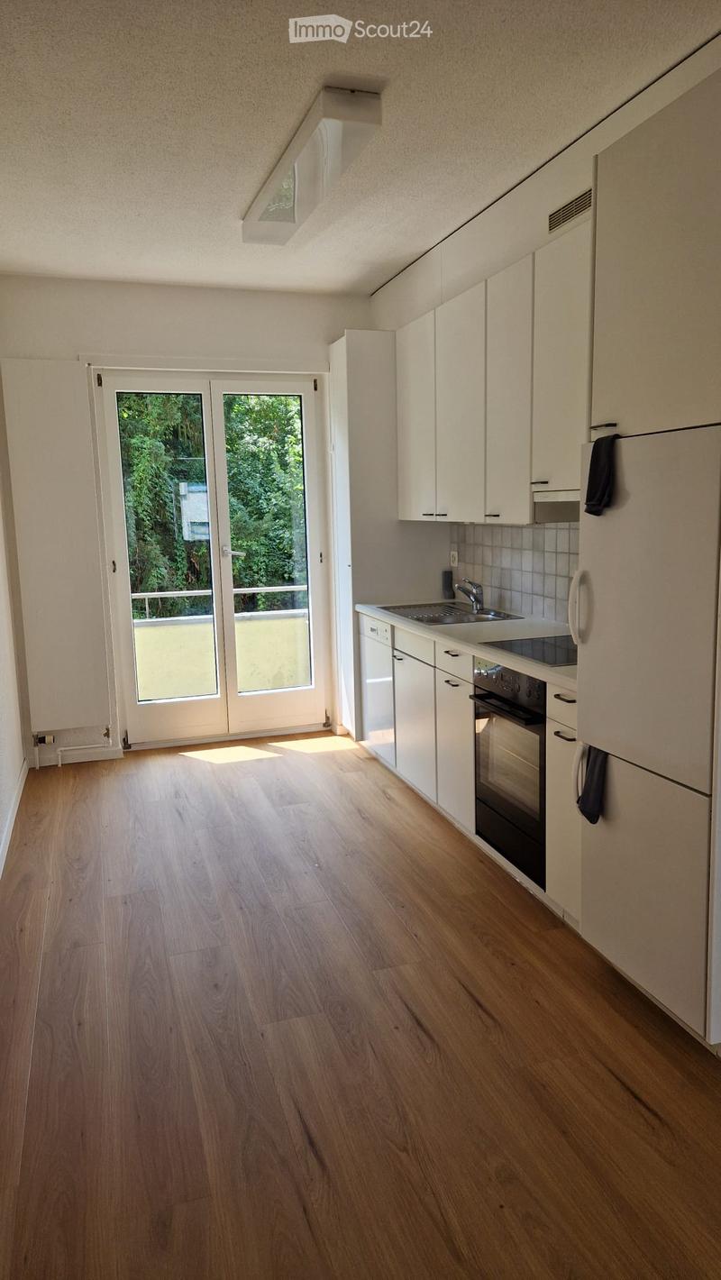 Modern kitchen with light wooden flooring, white cabinets, sink, and oven. Large sliding glass door offers a view of greenery outside.