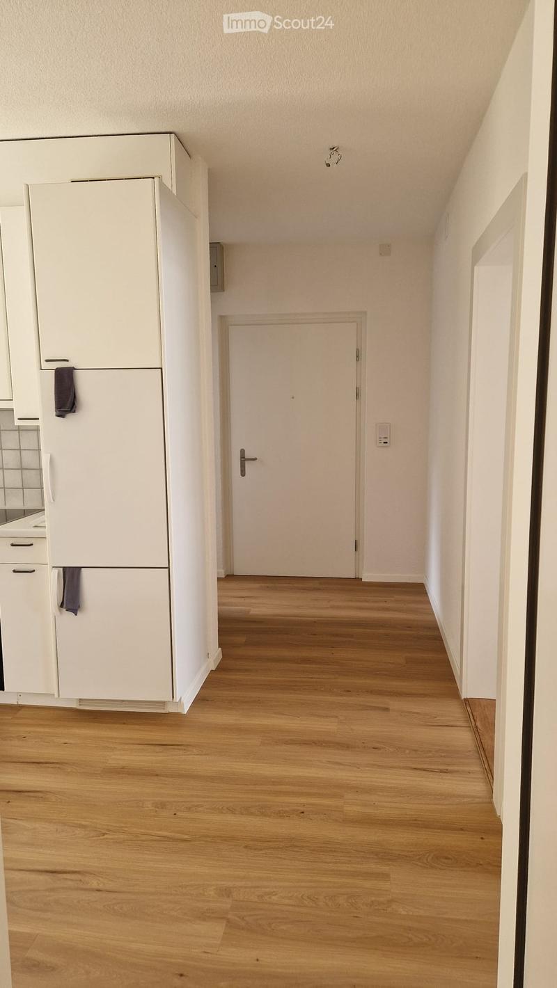 Empty hallway with wooden flooring, white walls, and a closed door at the end. A white cabinet and a refrigerator are visible on the left side.