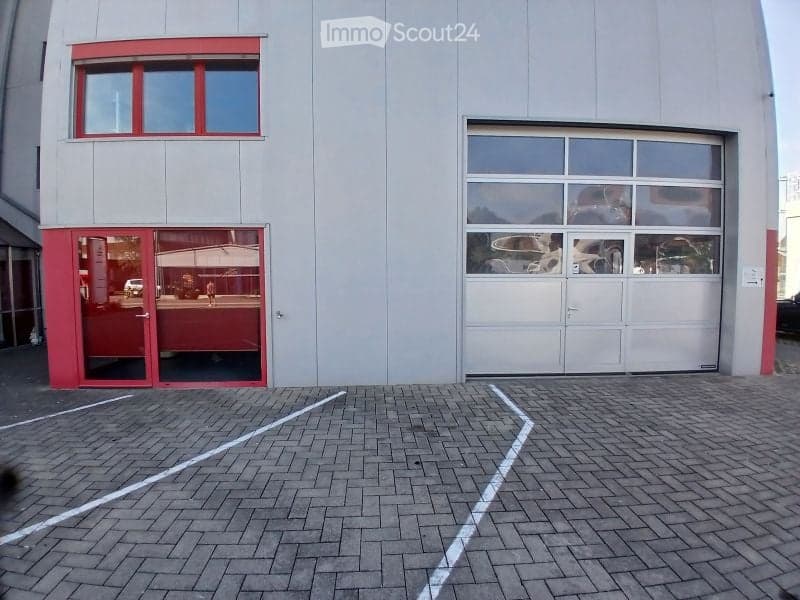 Modern building with a large garage door and a glass entrance. The facade is grey with red trim around the windows and doors. The ground is paved with bricks and marked parking spaces.