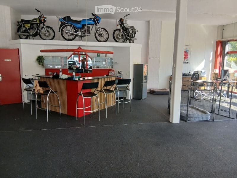 A modern reception area with a red counter, several stool chairs, a refrigerator, and motorcycles displayed on a shelf above the counter. There is also a fenced area in the corner.