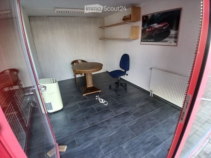 A small, furnished room with a round wooden table, two chairs, an office chair, a radiator, shelves, and a picture frame on the wall.