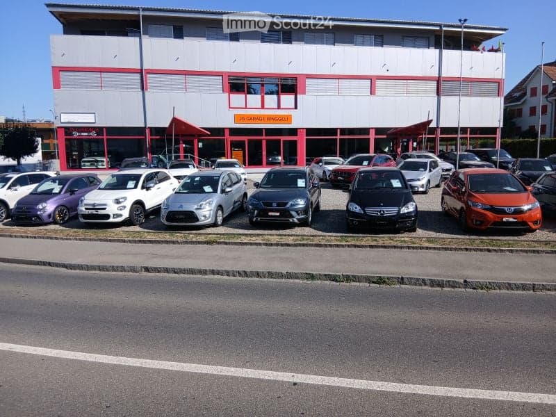 Modern two-story building with a red and gray facade, large windows, and a sign reading "Garage Binggeli". Several cars are parked in front of the building.