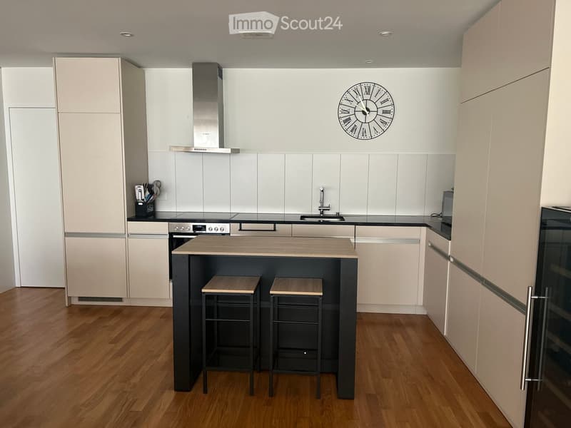 Modern kitchen with light-colored cabinets, a central island, two stools, a stove, sink, and a clock on the wall.