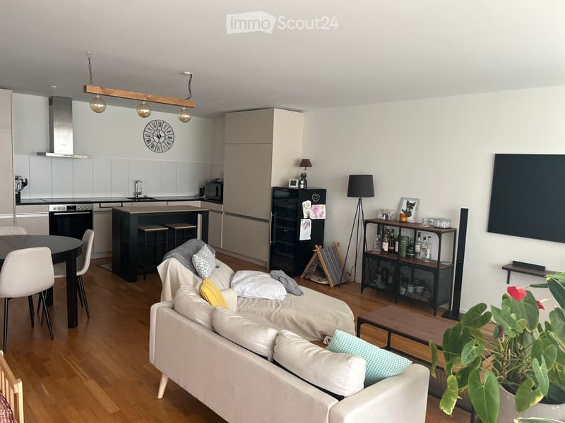 Modern living room with a white sofa, coffee table, kitchen area, and dining table. Features include a large TV, floor lamps, and a bottle rack.