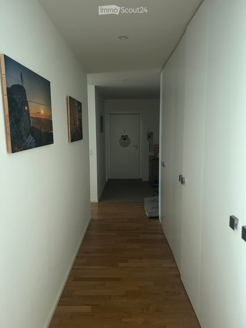 A narrow hallway with light wooden flooring, white walls, and two framed pictures on the left side. A closed door is visible at the end of the hallway, with a small decorative item hanging on the wall near the door.