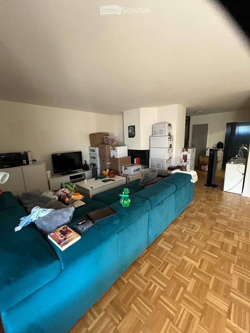 Living room with a teal sofa, coffee table, TV, laptop, and various items scattered around, including boxes and a refrigerator in the background.