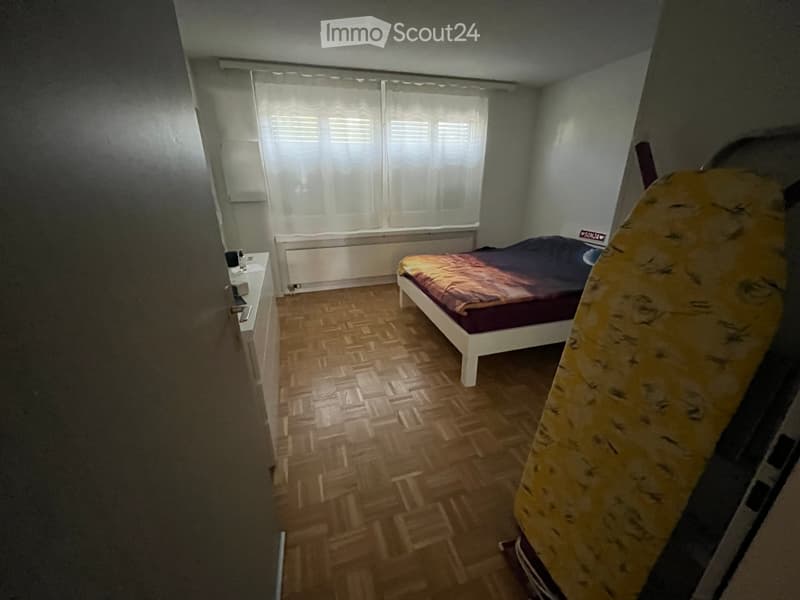 Bedroom with wooden parquet flooring, a single bed with a colorful duvet, a nightstand with a mirror, and a window with white curtains.