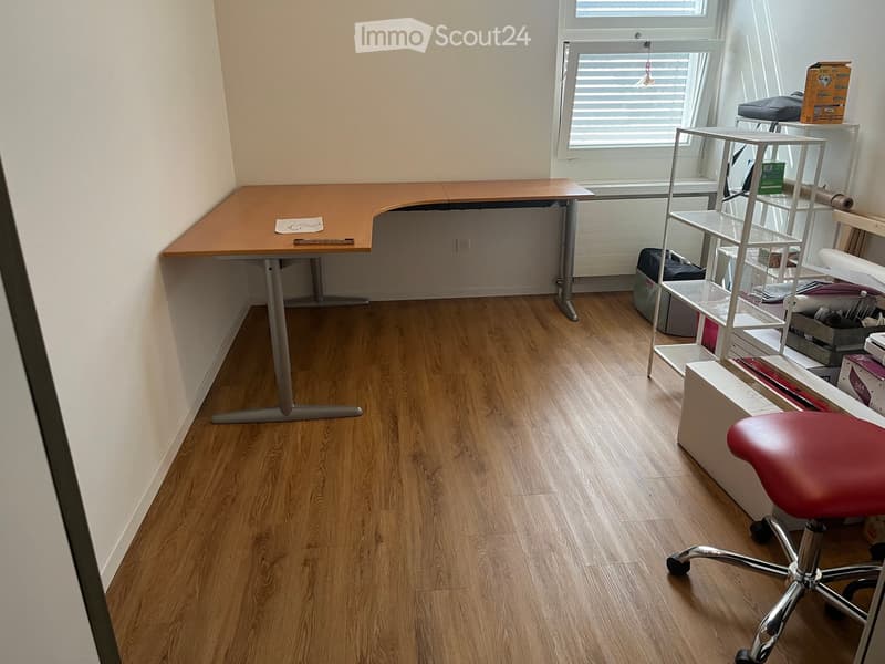 A furnished room with a wooden floor, a large desk, a red office chair, and a metal shelving unit with various items.