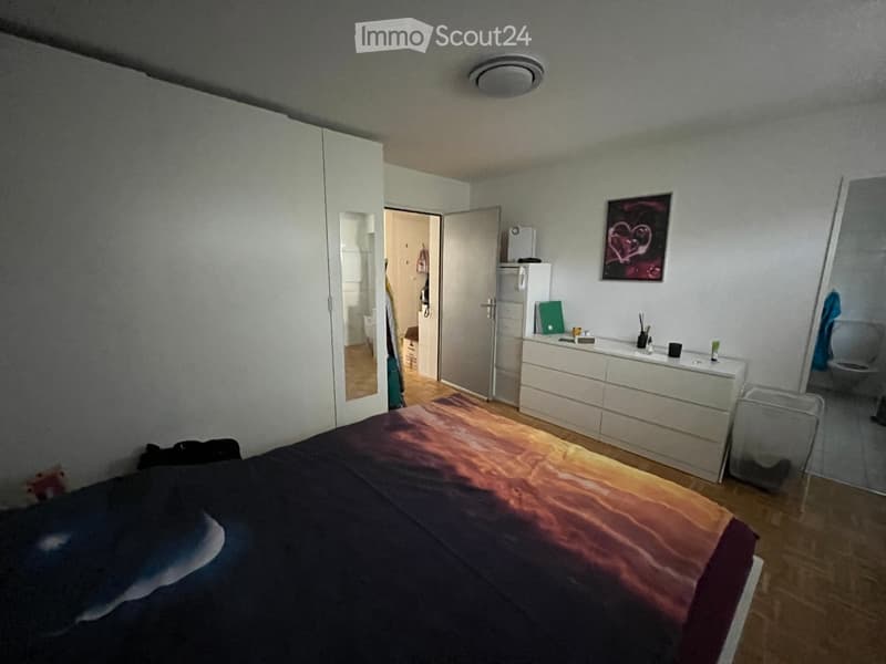 Bedroom with a bed, wardrobe, dresser, mirror, and a small fridge. The room has wooden flooring and white walls.