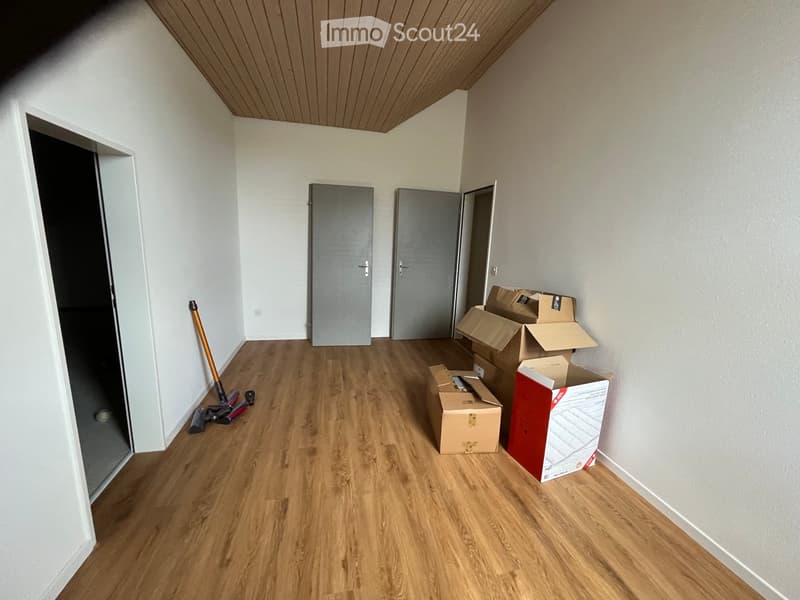 Empty room with wooden flooring, white walls, and several doors. Cardboard boxes and cleaning tools are present.