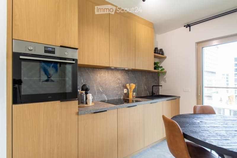 Modern kitchen with light wood cabinets, stainless steel oven, sink, and countertop. Includes a small dining area with a table and chairs, and a view of the city through large windows.