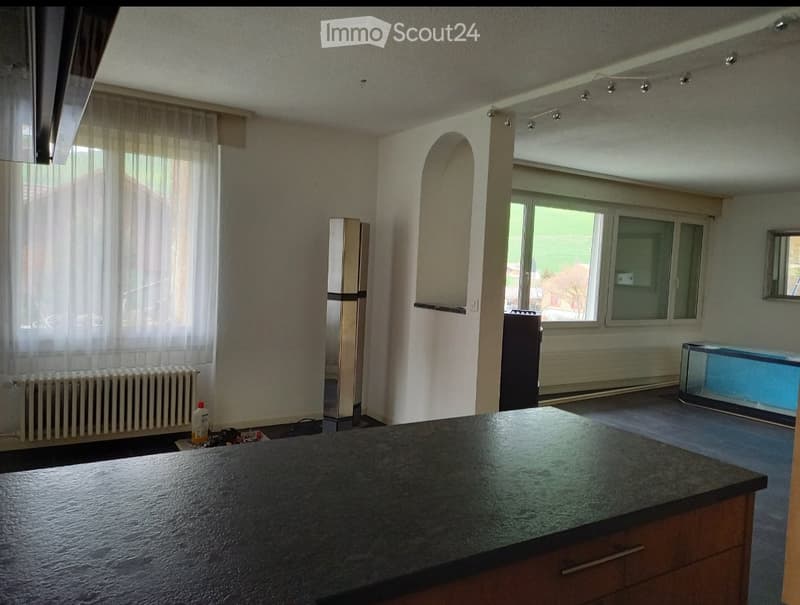 Empty kitchen with a dark countertop, wooden cabinets, radiator, and a refrigerator. Large windows with sheer curtains allow natural light to enter.