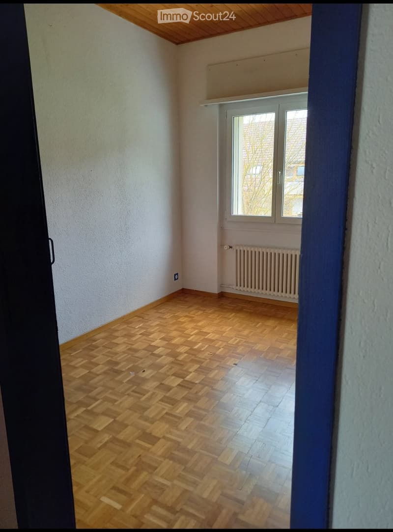 Empty room with wooden herringbone flooring, white walls, and a window with a radiator below it.