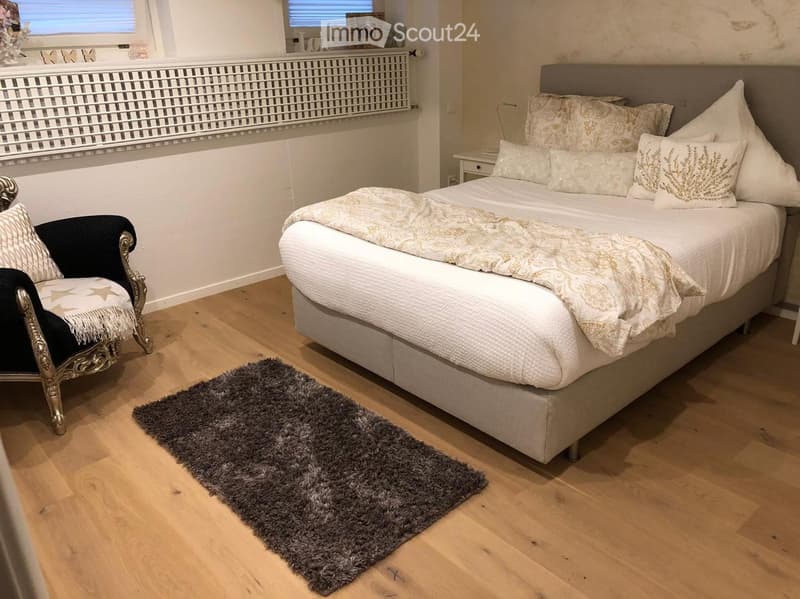 Cozy bedroom with a large bed, patterned bedding, a decorative chair, a nightstand, and a plush rug on a wooden floor.