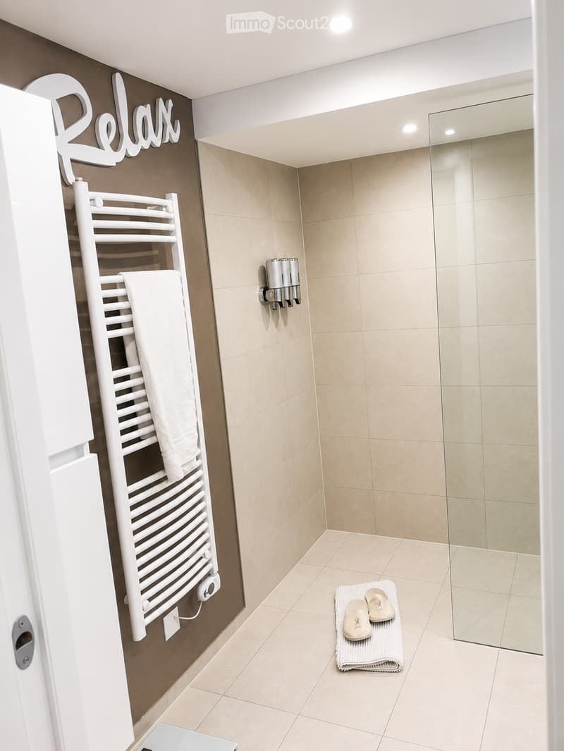Modern bathroom with beige tiles, a white towel rack with towels, a glass-enclosed shower, a soap dispenser, and a folded bathrobe with slippers on a towel.