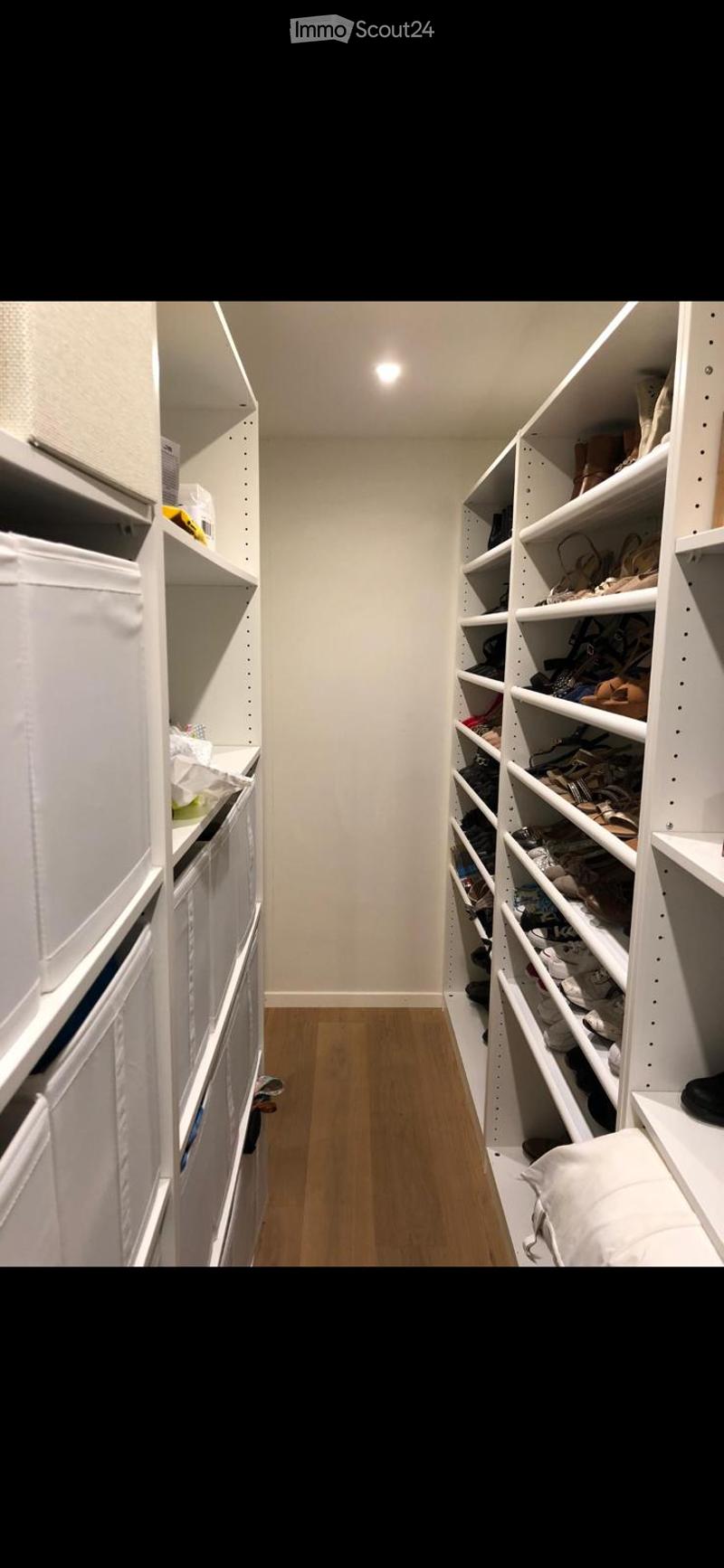 Spacious, well-lit closet with white built-in shelving units on both sides, containing neatly organized clothing, shoes, and storage boxes. The floor is light wooden, and the walls are white.