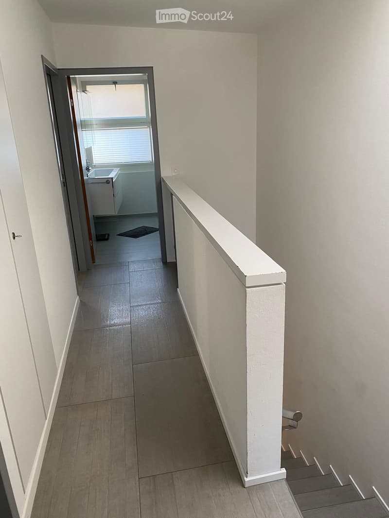 A narrow hallway with tiled flooring, white walls, and a staircase leading upwards. An open doorway leads to a small bathroom with a sink, toilet, and bathtub.