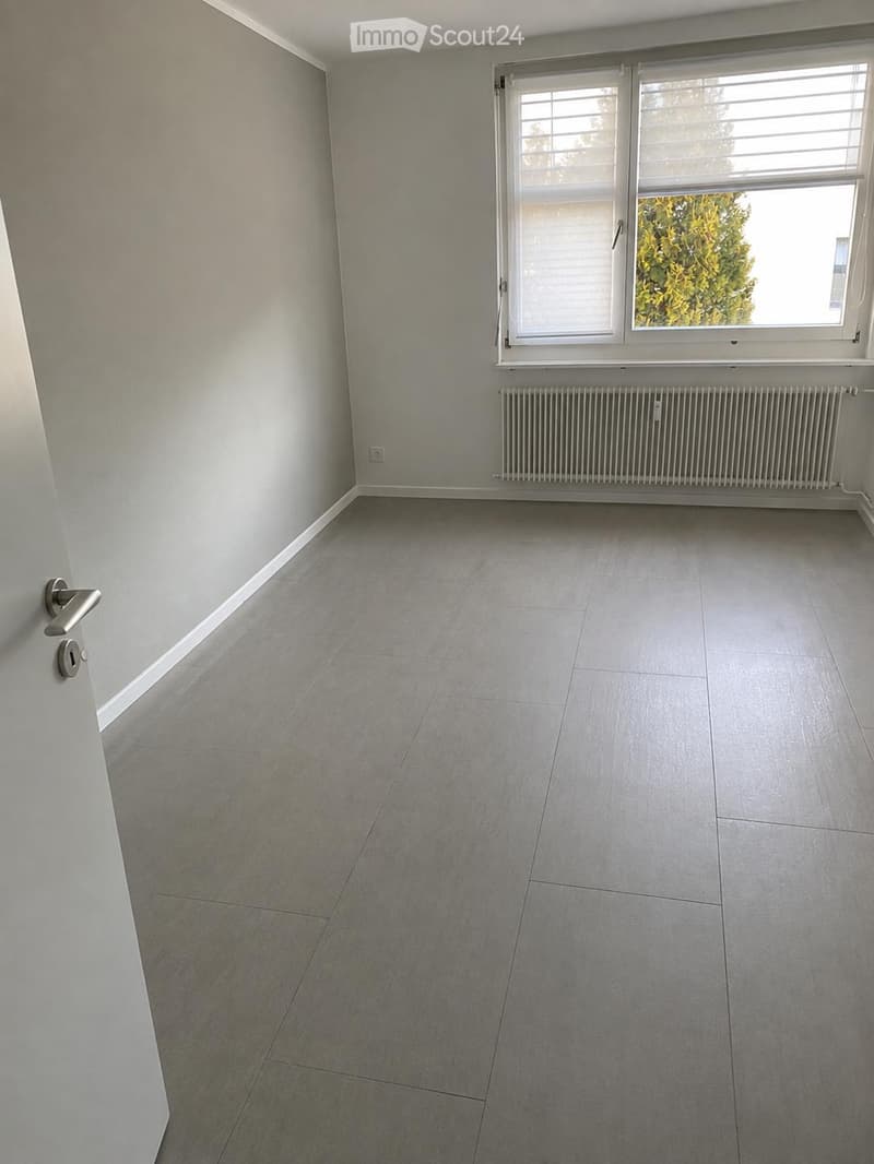 Empty room with light grey tiled flooring, white walls, a window with blinds, and a radiator below the window. The door handle is visible on the left side.