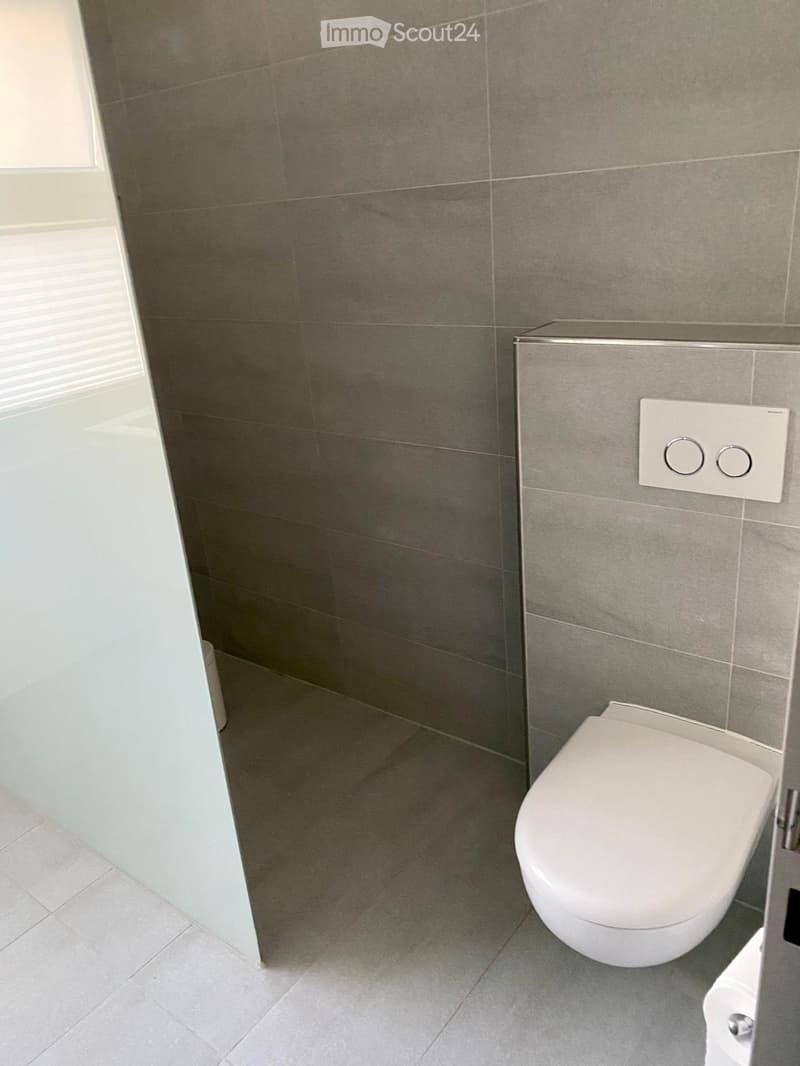 Modern bathroom with grey tiled walls and floor, white toilet, and a control panel on the wall.