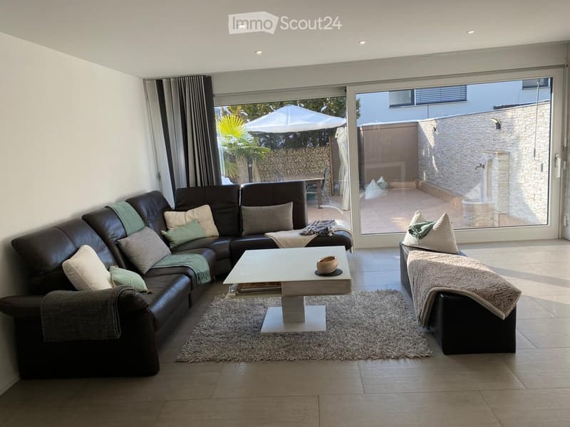 Modern living room with a dark leather sectional sofa, a white coffee table, and a shaggy rug. Large glass doors open to a balcony with outdoor furniture.