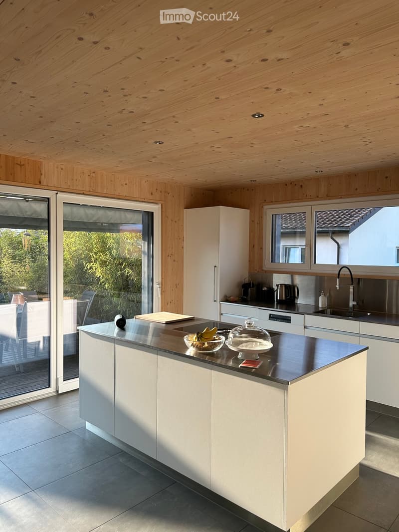 Modern kitchen with white cabinets, a central island, and a black countertop. The island features a bowl of fruit and a glass dome. The kitchen has a sleek design with a stainless steel sink and faucet, and a large window offers a view of the outdoor area