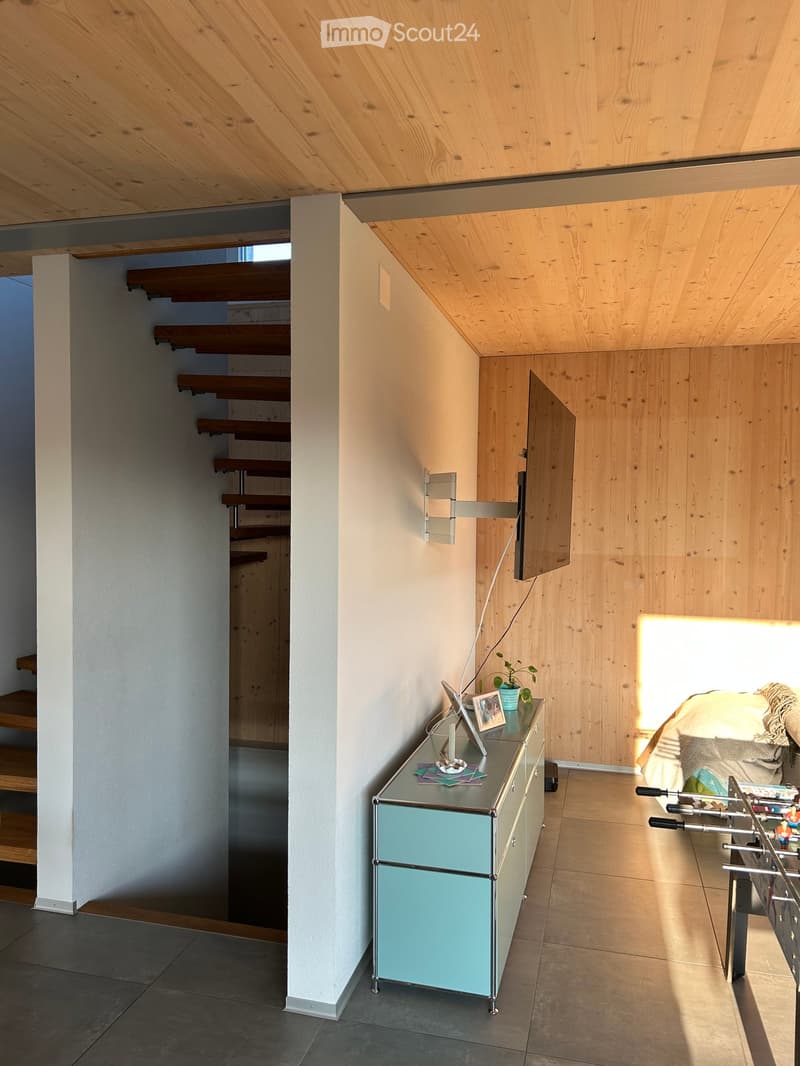Modern living room with wooden ceiling, grey tiled floor, and a staircase leading upwards. Features a glass-top table with a blue base, a mounted TV, and a small plant on the table.