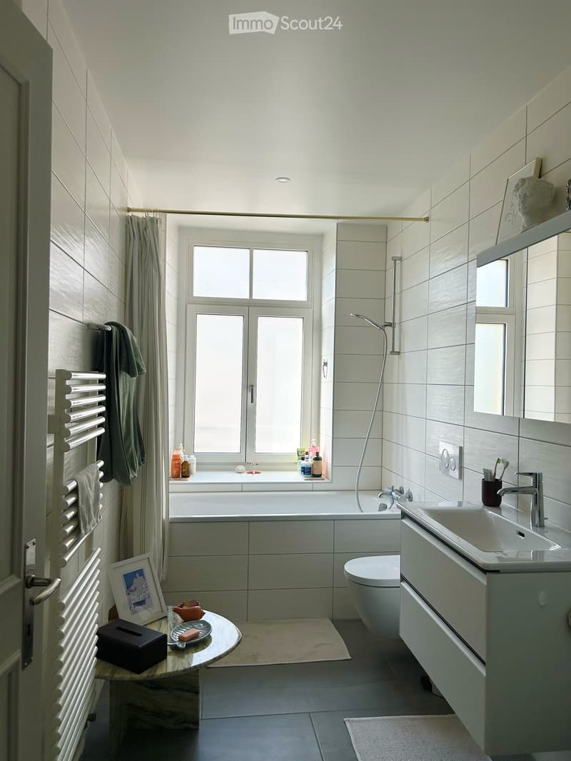 Modern bathroom with a bathtub, toilet, sink, and shower area. Features include a radiator, towels, a small table with a framed picture, and various bathroom accessories.