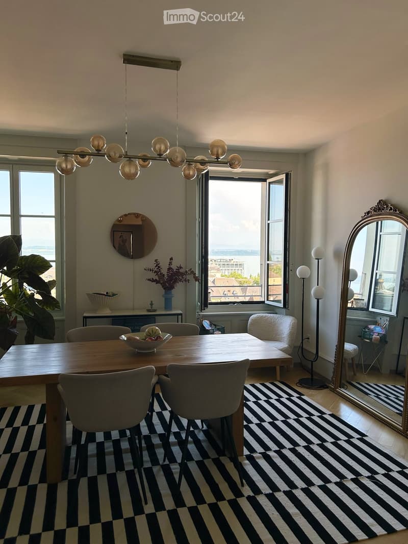 Modern dining area with a wooden table, chairs, a sofa, floor lamp, large windows offering a view, and decorative items including a mirror and plant.