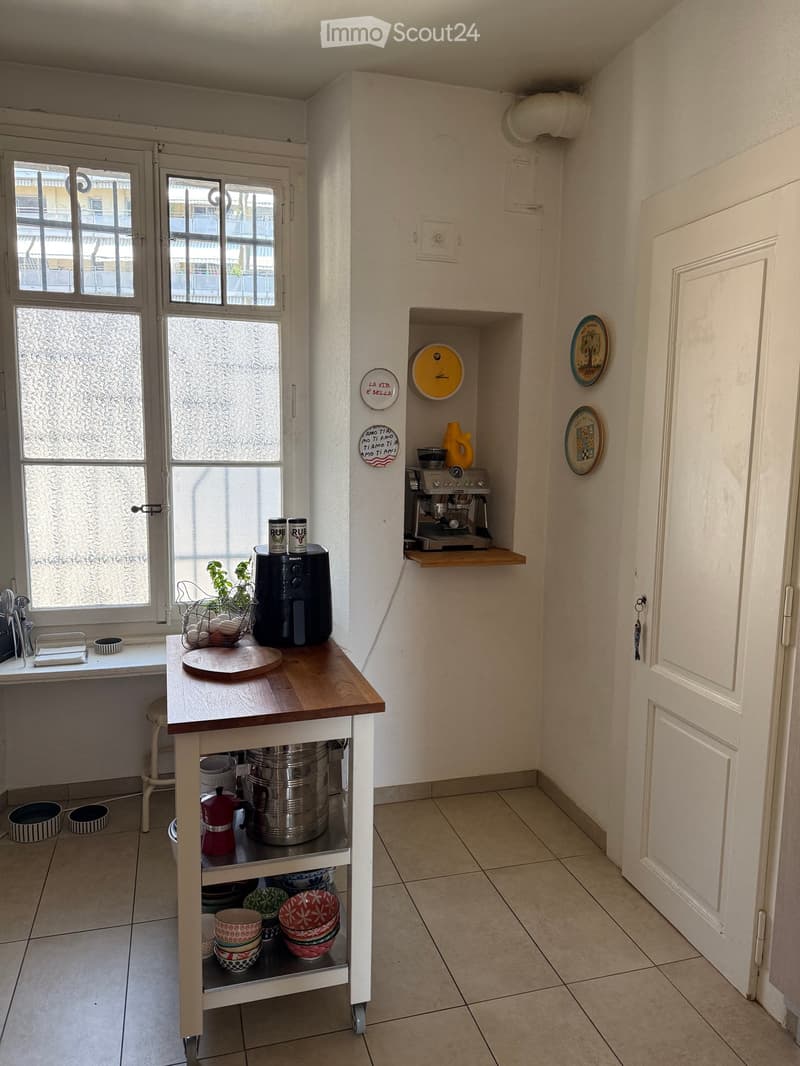 The kitchen features a wooden table with a stool, a coffee maker, an air fryer, and various kitchen items including bowls and cups. There is a built-in shelf with decorative plates and a small plant on the windowsill.