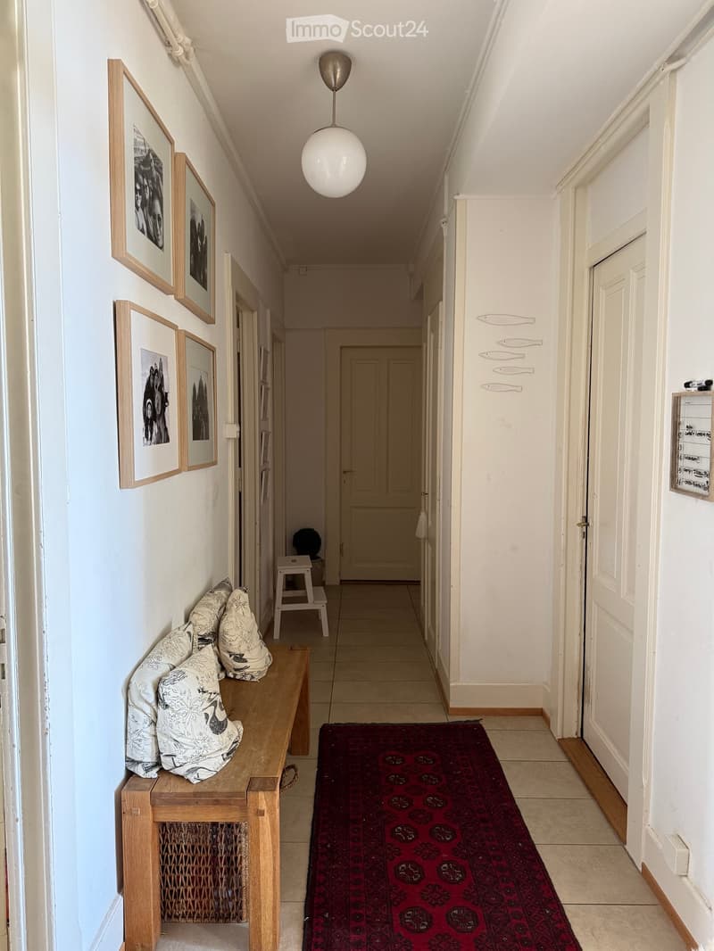 A well-lit hallway with white walls, wooden doors, a red runner rug, a wooden bench with patterned cushions, and framed artwork on the walls.