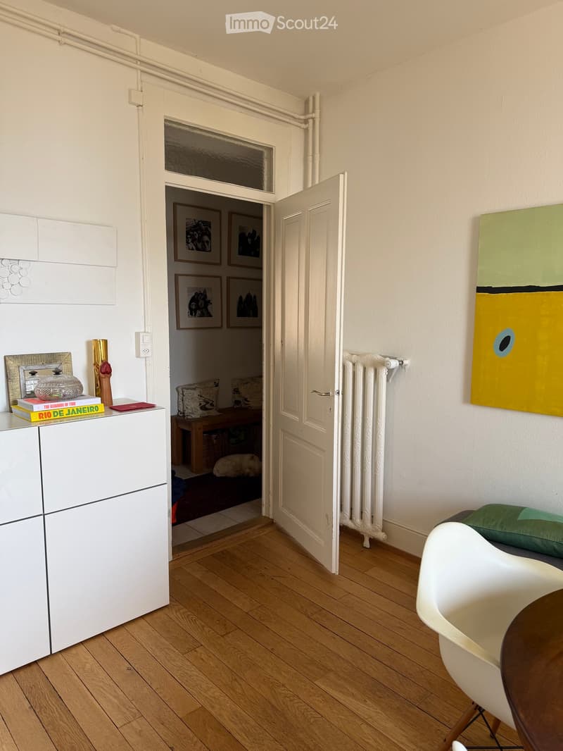 A modern, well-lit room with wooden flooring, white cabinetry, a white chair, a small table, a radiator, and an open doorway leading to another room with a bench and pillows.
