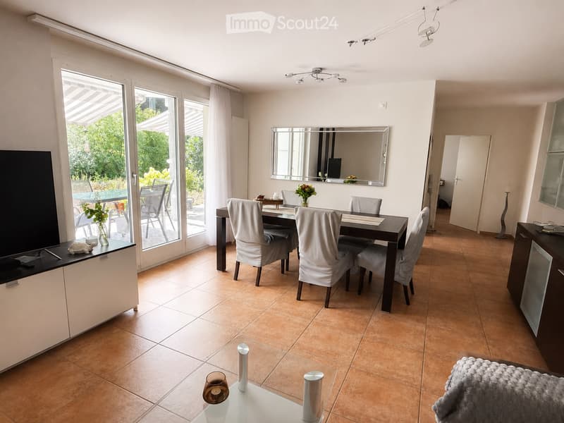 Bright, open-plan living and dining area with large windows and sliding doors leading to a patio. Features a dining table with six chairs, a TV on a stand, a mirror on the wall, and a small coffee table with decorative items.