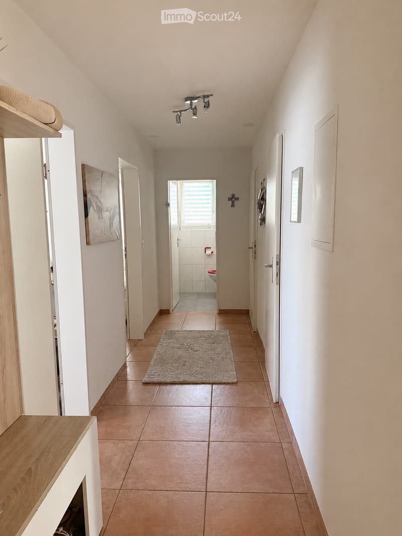 A clean, well-lit hallway with beige tiled flooring, white walls, and a small rug in the center. A painting is visible on the left wall, and a bathroom is visible at the end of the hallway.