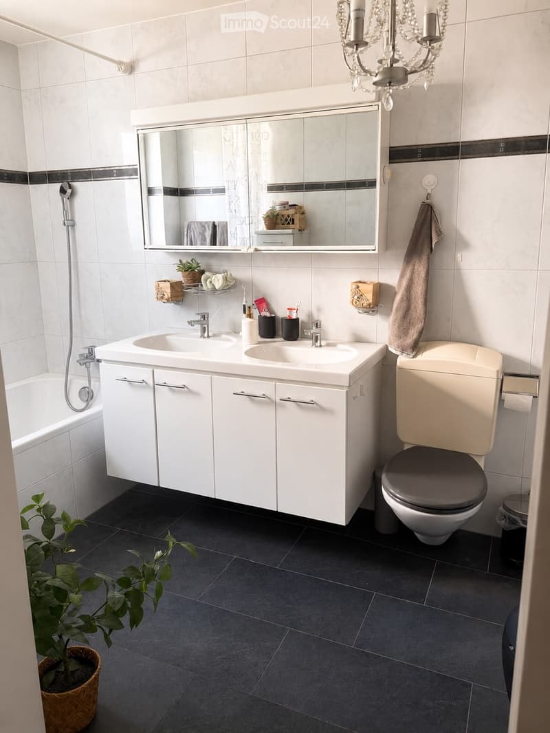 Modern bathroom with white tiles, dark grey floor, double sink vanity, toilet, bathtub, large mirror, and decorative items including a plant and a chandelier.
