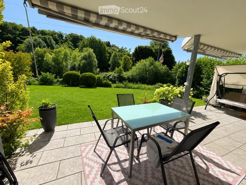 Outdoor patio with a glass table, chairs, and a swing bed, surrounded by a well-maintained lawn and trees.