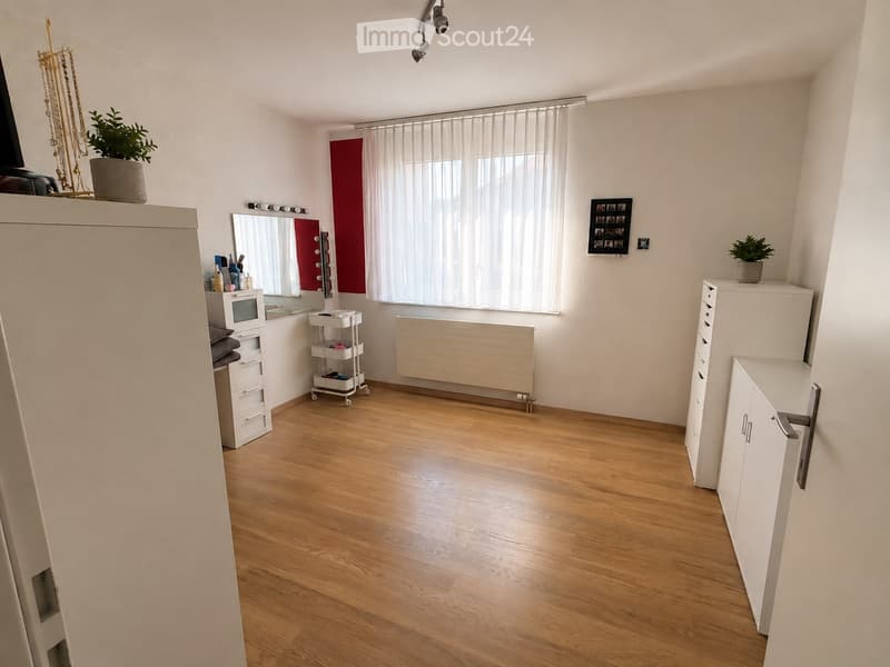A well-lit room with wooden flooring, white walls, and large windows covered with vertical blinds. Features include a mirror, cabinets, a small cart, and decorative plants.