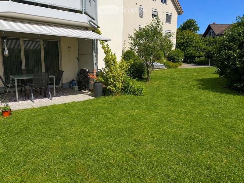 Well-maintained lawn with a patio area featuring a table, chairs, and potted plants, adjacent to a modern house with a covered balcony.