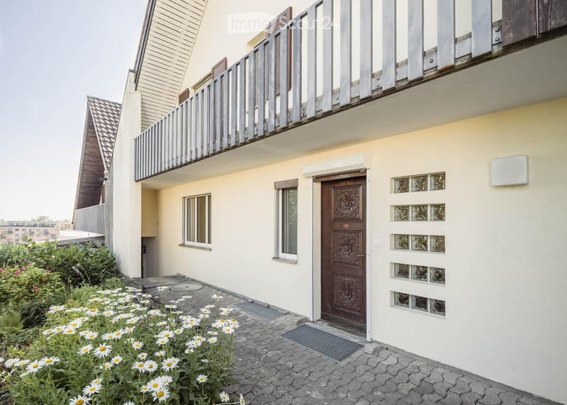 Two-story house with a balcony, beige exterior, wooden front door, and a small garden with white flowers.