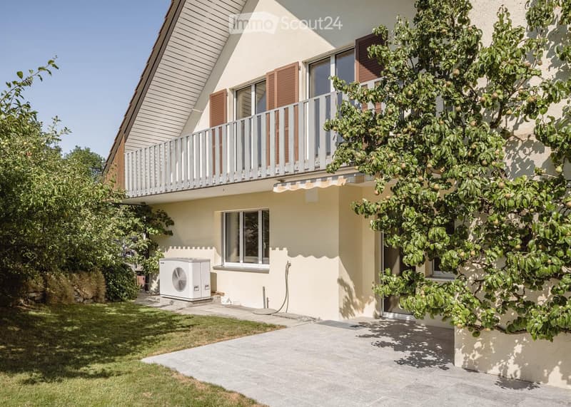 Two-story house with a balcony, beige exterior, wooden shutters, and a small garden area with a tree and air conditioning unit.