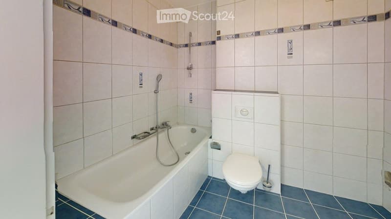 Bathroom with a bathtub, toilet, showerhead, and faucet. The walls are tiled with a decorative border, and the floor is blue tiled.