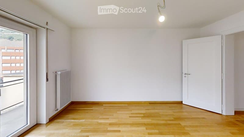 Empty room with wooden flooring, white walls, a radiator, and a door leading to another space.