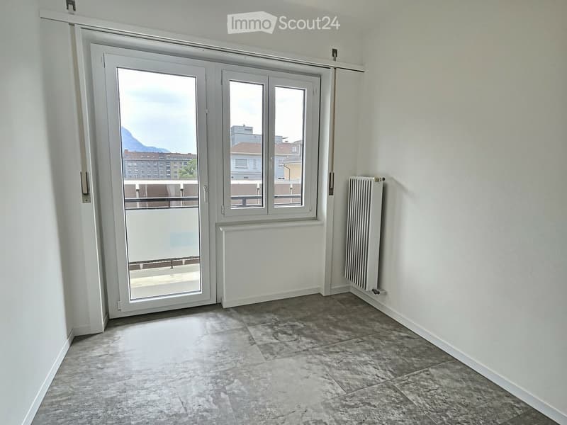 Empty room with grey tiled floor, white walls, large sliding glass door leading to a balcony, and radiators on both sides of the door.