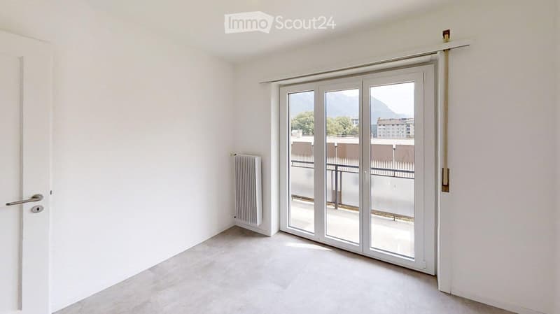 Empty room with white walls, light grey tiled floor, large sliding glass door leading to a balcony, radiator on the wall, and a door with a silver handle.